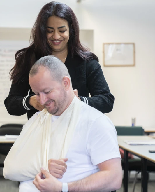 One person helps another put on an arm sling in a classroom, practicing first aid skills with care and teamwork One person helps another put on an arm sling in a classroom, practicing first aid skills with care and teamwork